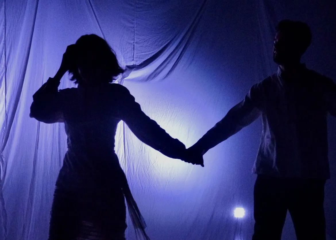 Two silhouetted figures joining hands against a blue-lit sheet.