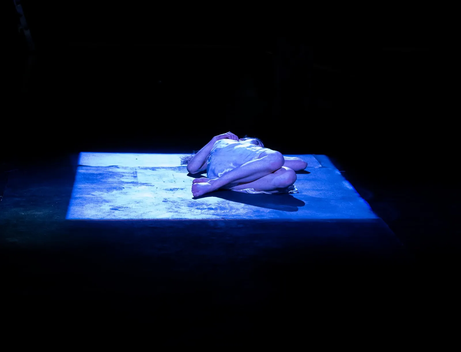 Eurydice curled alone in a lit square on a black stage.