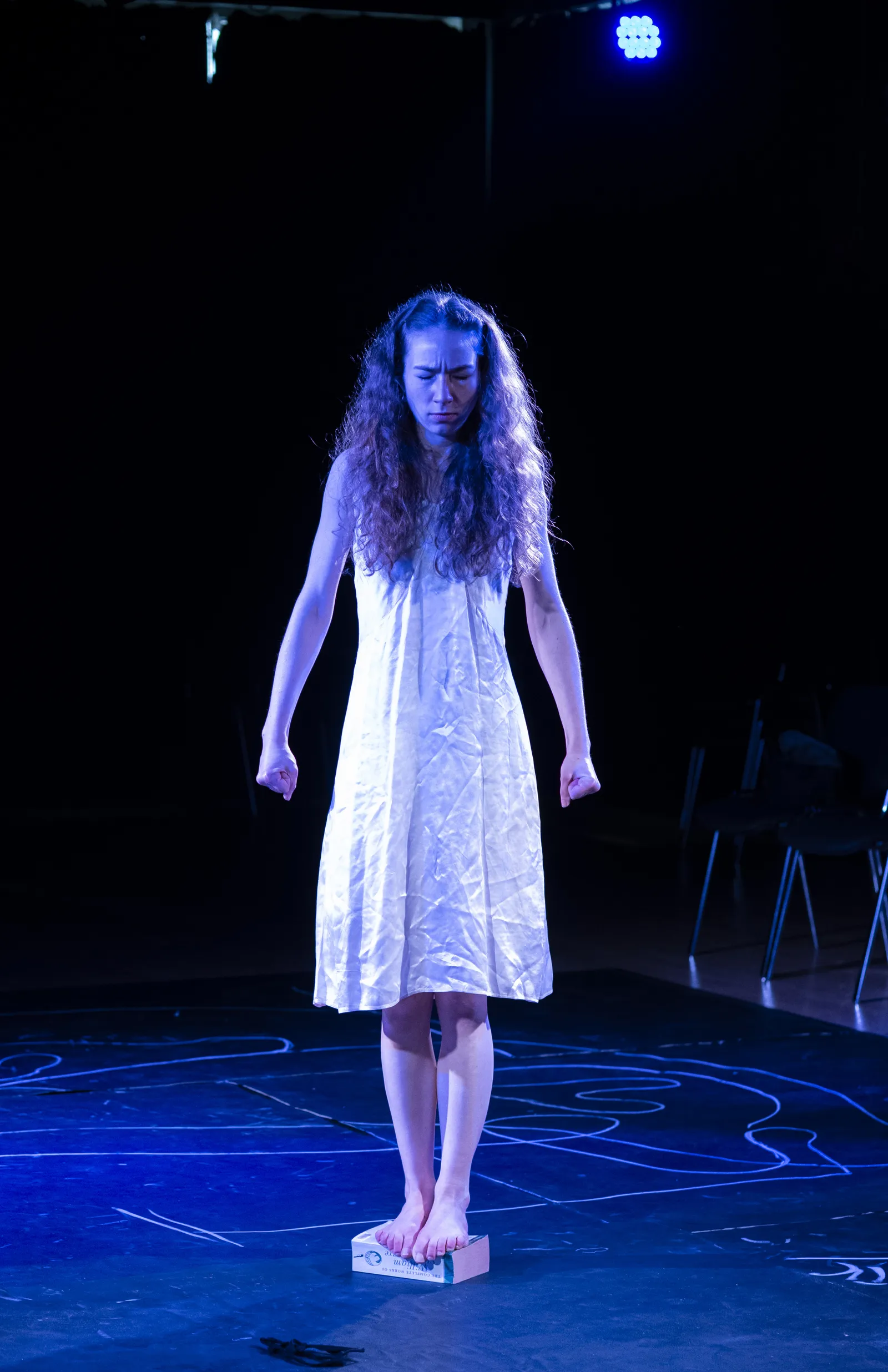 Eurydice in a white dress standing on a small block in blue light.